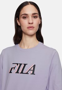 Violetti huppari, jossa on ribbauskaulus, ja edessä on suuri musta ja punainen "FILA"-logo. Pehmeä, tekstuurinen kangas.