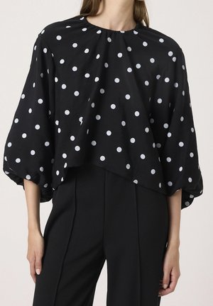 Kobieta ubrana w luźną czarną bluzkę w białe kropki z balonowymi rękawami o długości do połowy ramienia, zestawioną z wysokim stanem czarnych spodni.