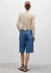 Haut à manches longues en dentelle crème avec des motifs élaborés, associé à un short Bermuda en denim bleu et des sandales noires à lanières.