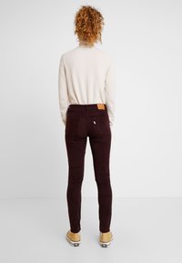 Pantalones skinnies de pana burdeos con una etiqueta de cuero marrón en la cinturilla trasera, combinados con un suéter color crema claro y zapatos amarillos.