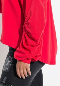 Rotes Langarmoberteil mit gerafftem Detail an den Bündchen, kombiniert mit schwarzen verwaschenen Jeans. Der Stoff wirkt glatt und leicht.