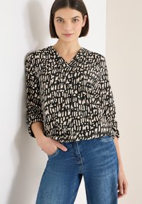 Sort og cremefarvet mønstret bluse med V-udskæring og opsmøgede ærmer, båret med blå jeans. Stoffet ser blødt ud med en glat tekstur.