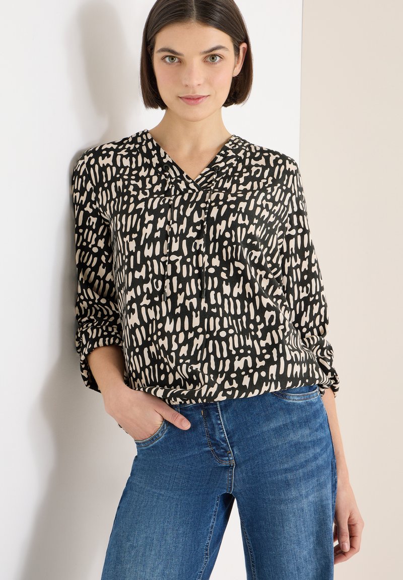 Sort og cremefarvet mønstret bluse med V-udskæring og opsmøgede ærmer, båret med blå jeans. Stoffet ser blødt ud med en glat tekstur.