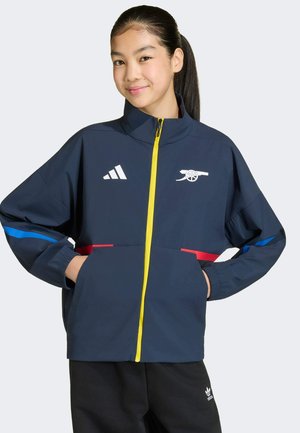 Giacca navy con cerniera, colletto alto, cerniera gialla, accenti laterali rossi e blu, logo Adidas e grafica Arsenal sul petto.