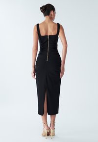 Vestido negro ajustado con tirantes anchos, espalda cuadrada y cremallera trasera. Presenta detalles fruncidos y una abertura alta en el muslo. Modelo con tacones de tiras.