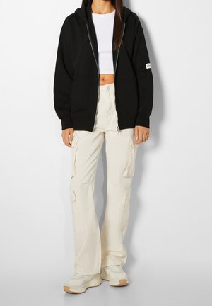 Kvinde iført en sort hættetrøje med lynlås, hvid cropped top, cremefarvede cargobukser og hvide sneakers står foran en ensfarvet baggrund.
