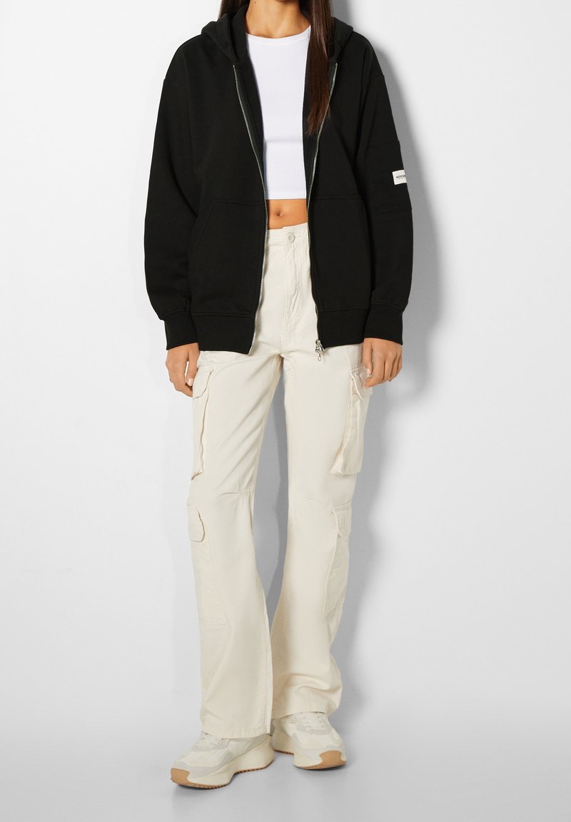 Femme portant un sweat à capuche noir zippé, un top court blanc, un pantalon cargo crème et des baskets blanches, debout devant un fond uni.