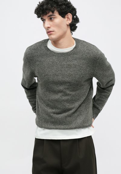 Šedý texturovaný pletený svetr s kulatým výstřihem, na němž je vrstvená bílá košile. Model má na sobě tmavé, volné kalhoty.
