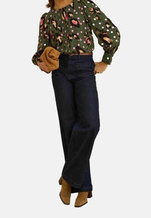 Mujer que lleva jeans anchos oscuros, blusa verde con estampado rosa y de leopardo, sosteniendo un bolso de mano marrón claro y botas marrones con tacón.