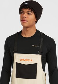 Overoles de pesca beige de O'Neill con bolsillo negro, tirantes ajustables y camiseta de manga larga negra y ligera. Gorro negro con logotipo.