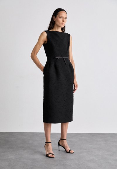 Svart, ärmlös klänning i strukturerad tyg, med rak silhuett, dolda fickor och ett tunt midjebälte. Svarta högklackade sandaler.