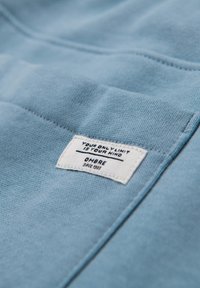 Lyseblåt stof med en lille hvid rektangulær etiket syet på, der viser teksten: "DIN ENESTE BEGRÆNSNING ER DIT SIND" og "OMBRE siden 1989".