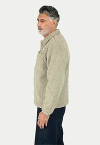 Giacca in pile beige con finitura testurizzata, colletto mezza zip e tasca sul petto, abbinata a jeans scuri.
