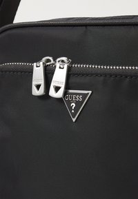 Borsa nera in nylon con doppie cerniere argentate dotate di tiranti triangolari e un badge logato triangolare con la scritta "GUESS USA" in rilievo.