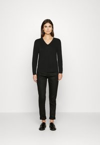 Schwarzer V-Ausschnitt-Pullover mit langen Ärmeln, kombiniert mit schwarzen Slim-Fit-Hosen. Modell trägt schwarze Slipper und steht vor einem neutralen Hintergrund.