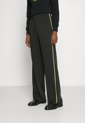 Μαύρες παντελόνες με φαρδιά πόδια, με λεία υφή και νεον κίτρινες ρίγες στο πλάι. Συνοδευόμενες από μαύρα παπούτσια με διακριτικά τρουκς.