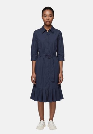 Woman wearing a navy blue buttoned dress with a belt at the waist and ruffled hem, paired with white sneakers, standing on white background.
