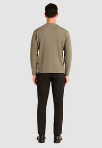 Maglione verde oliva con scollatura rotonda, maniche lunghe, indossato con pantaloni neri e scarpe scure, visto da dietro su uno sfondo grigio.