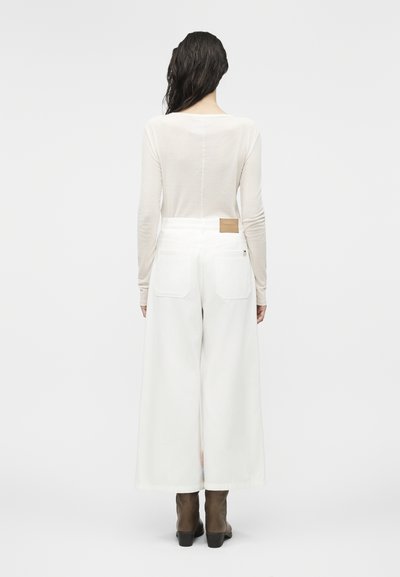 Γυναίκα με υγρά σκοτεινά μαλλιά φοράει διαφανή μπλούζα με μακριά μανίκια, άσπρο παντελόνι φαρδιάς γραμμής με πίσω τσέπες και καφέ μποτάκια αστραγάλου, με την πλάτη προς τα εμπρός.