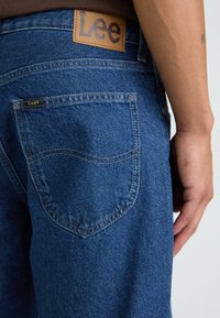 Jeans din denim de culoare albastră medie cu o textură netedă, având un ecuson din piele pe talie și o etichetă mică pe buzunarul din spate.