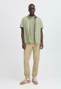 Chemise à manches courtes vert clair avec un bouton sur un t-shirt assorti, pantalon beige fuselé et baskets marron avec des accents blancs.