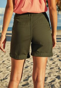 Olijfgroene katoenen korte broek met omgeslagen zoom en achterzakken, gecombineerd met een roze top. De achtergrond toont zand en strand.