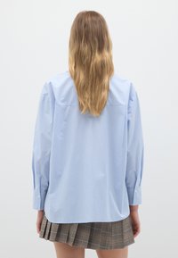 Camicia a righe azzurro chiaro con maniche lunghe e polsi bottonati, caratterizzata da una vestibilità comoda e dettagli di plissettatura sul retro, abbinata a una gonna a quadretti.