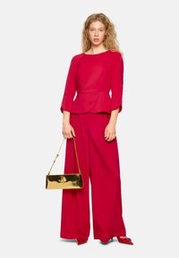 Rotes Peplum-Top und weite Hose aus glattem Stoff. Das Modell hält eine goldene metallische Clutch mit einem dekorativen Verschluss.