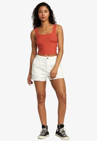 Rust-colored ribbed crop top with wide straps, paired with high-waisted white corduroy shorts and black high-top sneakers.
