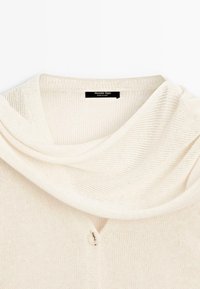 Krémový pletený svetr s volně splývajícím výstřihem a knoflíkem vpředu. Značka "Massimo Dutti" viditelná uvnitř límce.