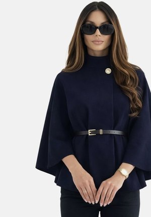 Femme aux longs cheveux ondulés portant des lunettes de soleil noires, une cape bleu marine foncé ceinturée avec un seul bouton doré, et une montre en or au poignet gauche.
