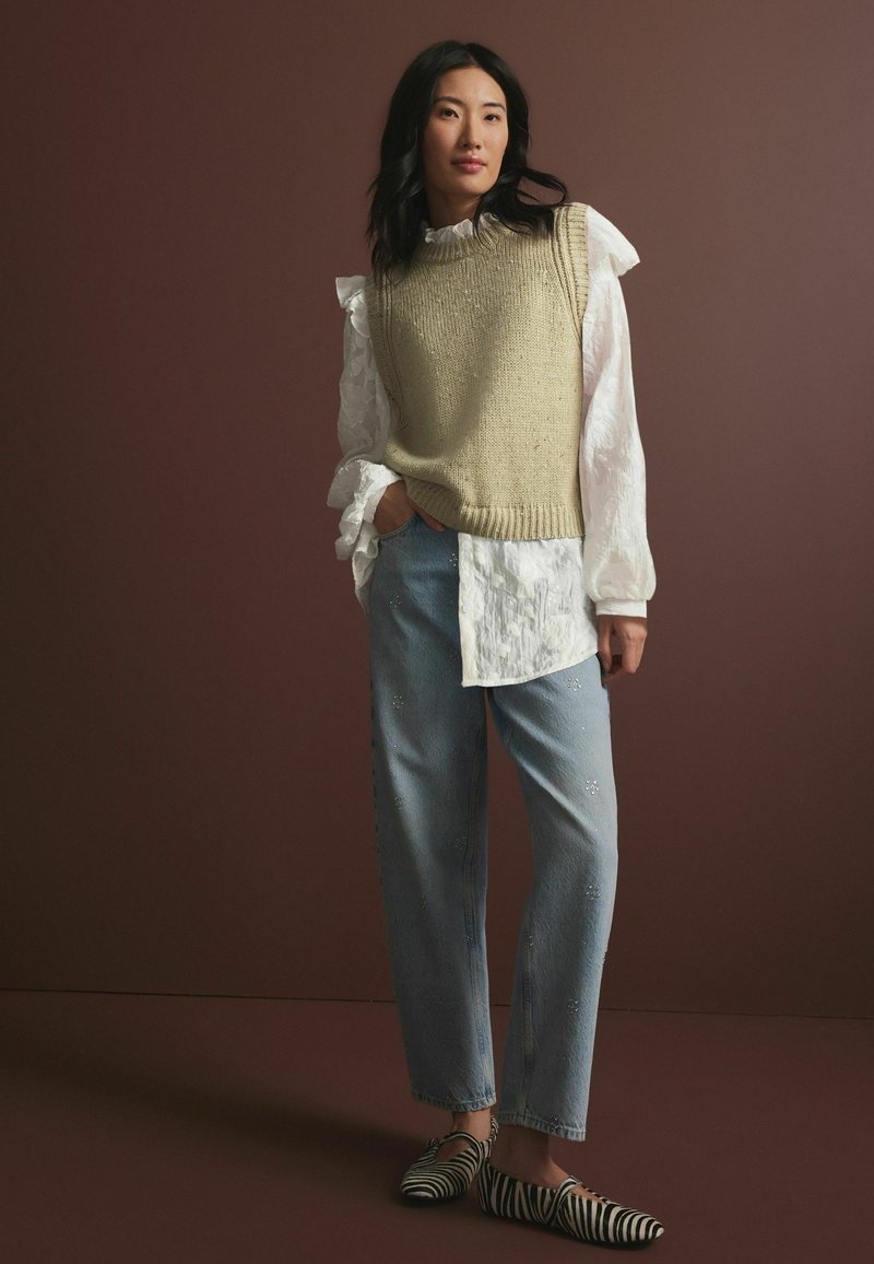 Strik beige ærmeløs sweater over en hvid bluse med rynker, parret med lyseblå jeans med dekorative detaljer og sort-hvide stribede sko.