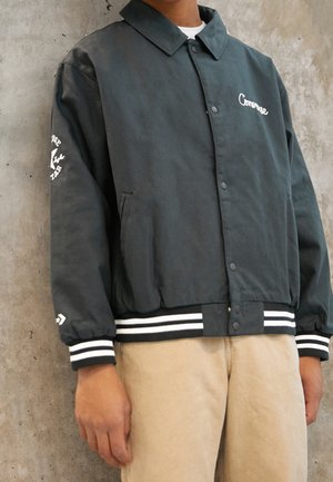 Personne portant une veste noire Converse à boutons avec un logo blanc sur la poitrine et la manche, des poignets rayés et un pantalon beige devant un mur en béton.