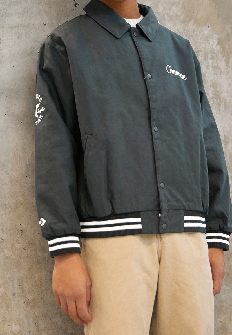 Personne portant une veste noire Converse à boutons avec un logo blanc sur la poitrine et la manche, des poignets rayés et un pantalon beige devant un mur en béton.