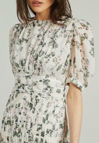 Blumenkleid mit Puffärmeln, das ein plissiertes Design aufweist. Weicher Stoff in Creme mit grünen Blättern und rosa Blumen. Taillierte Details.