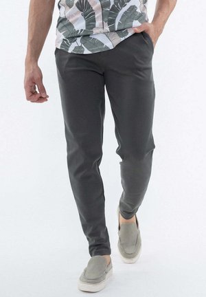 Homme portant un pantalon slim foncé, une chemise à motifs et des chaussures grises claires faciles à enfiler, marchant avec une main dans la poche.