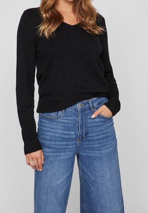 Vrouw met hand in de zak, gekleed in een zwarte V-hals trui met lange mouwen en een hooggesneden blauwe spijkerbroek, tegen een effen lichte achtergrond.