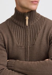 Maglione marrone a maglia con colletto alto, caratterizzato da motivi a coste e una zip frontale. Il tessuto appare texturizzato e caldo.