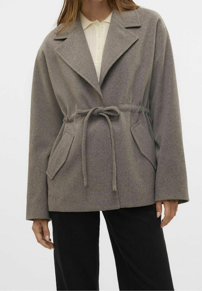 Grijze jas van een wolmix met een getailleerde taille, ingezette revers, twee voorzakken en een gestructureerde afwerking. Eronder een crème overhemd met knopen.