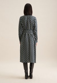 Sukienka z długim rękawem w ciemnoniebieskim kolorze z kremowym abstrakcyjnym wzorem w łańcuch, zaciśnięta w talii pasującym paskiem, w połączeniu z wysokimi czarnymi botkami.