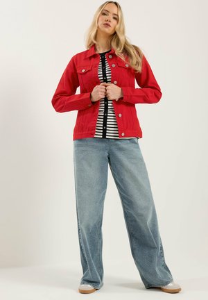 Kobieta o długich blond włosach, ubrana w czerwony zapinany na guziki żakiet, czarno-biały pasiasty top, szerokie niebieskie dżinsy oraz beżowe tenisówki, stoi prosto.
