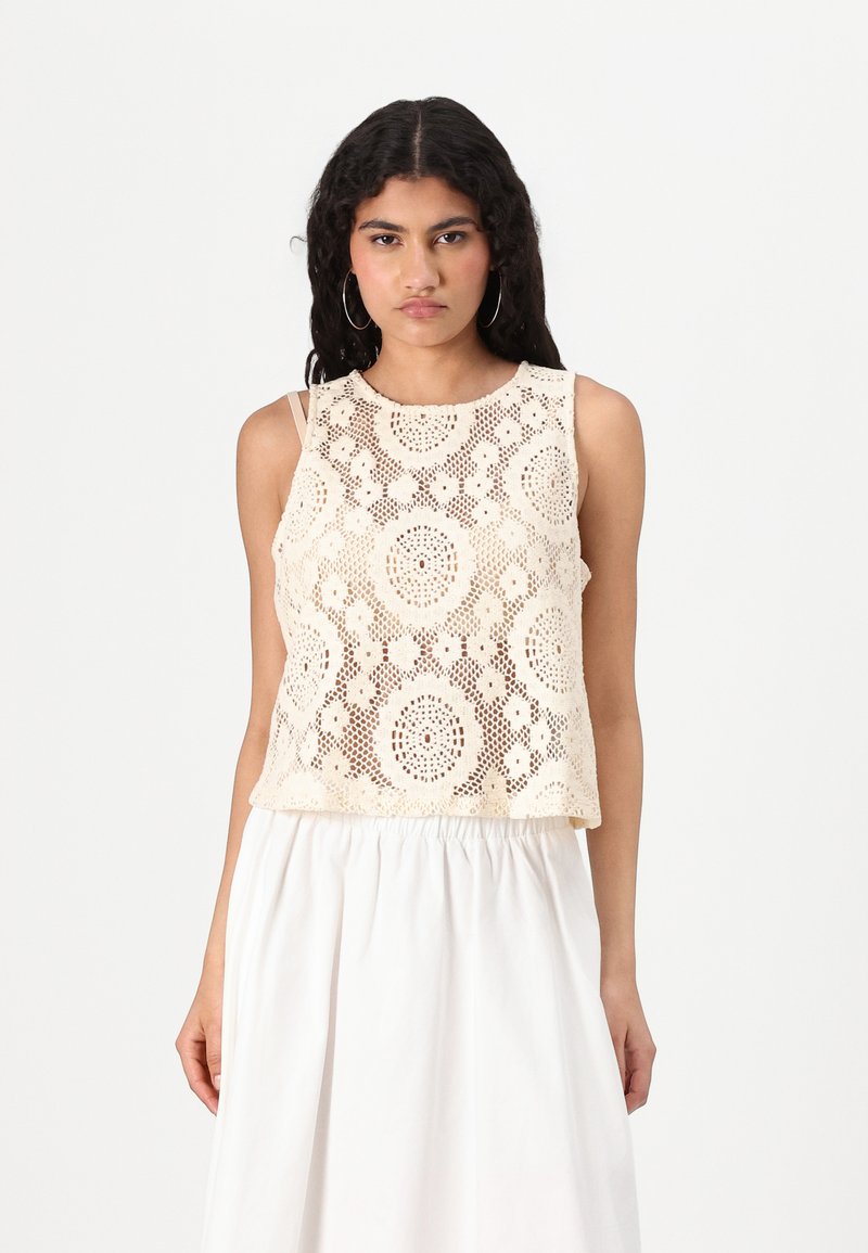 Blonde lace crop top i kremfarge, med blomster mønster og rund hals. Komplementert av tynne stropper og parret med et hvitt skjørt.