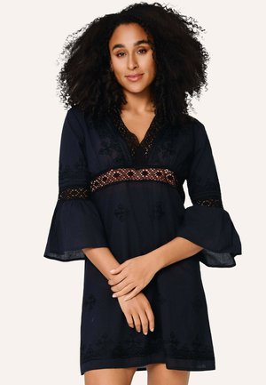 Femme aux cheveux bouclés portant une robe noire à larges manches et détails en dentelle, debout les mains croisées devant elle.