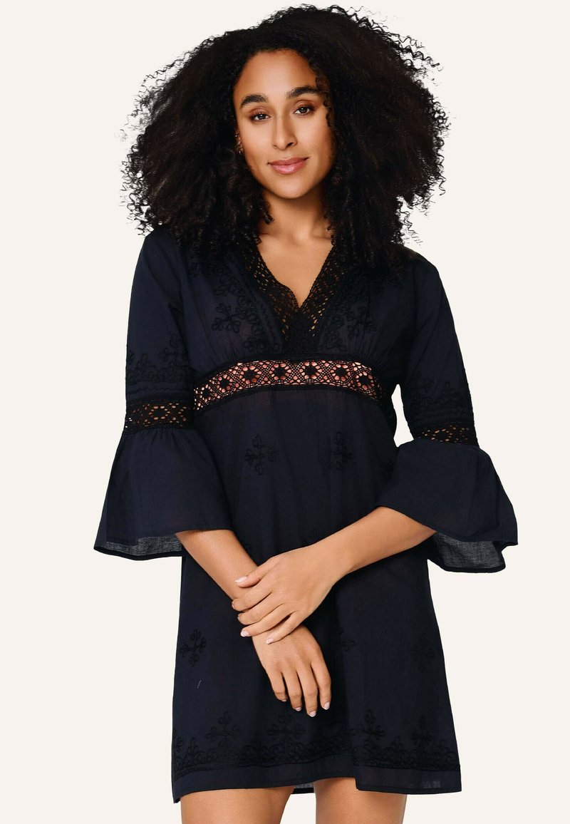 Kvinde med krøllet hår iført en sort kjole med vide ærmer og blonder, står med armene krydset foran sig.