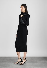 Vestido negro de manga larga con un ajuste ceñido al cuerpo y largo hasta la pantorrilla. Lleva tacones negros de tiras y un bolso negro colgado de un hombro.