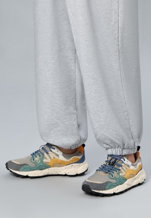 Person trägt graue Sweatpants und mehrfarbige Sneakers mit blauen Schnürsenkeln und gesprenkelten weißen Sohlen, steht auf einem grauen Boden.