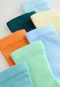 Sept paires de chaussettes côtelées pliées en bleu pastel, vert, orange, jaune, sarcelle et bleu clair, disposées en diagonale sur une surface neutre.