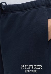 Σκούρο μπλε φόρμα με ελαστική μέση και άσπρο κείμενο «HILFIGER EST. 1985» κοντά στην τσέπη, το χέρι μερικώς ορατό στην τσέπη.