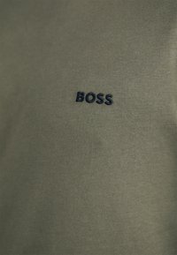 Alyvuogių spalvos medvilnės marškinėliai, kurių kairėje krūtinės pusėje išsiuvinėtas mažas tamsiai mėlynas "BOSS" logotipas. Lygus paviršius, kasdieniškas dizainas.