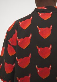 Tessuto nero con cuori rossi ripetuti caratterizzati da due piccole corna da diavolo indossato da una persona, visibile dalla parte posteriore e dalla spalla.